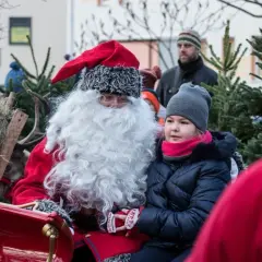 Święty Mikołaj w saniach z dziewczynką, która z zaciekawieniem patrzy przed siebie. W tle widać renifera i choinki.