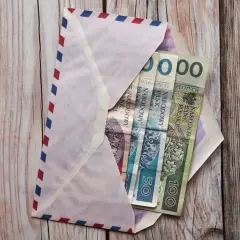 Koperta z polskimi banknotami (50 zł, 100 zł) na drewnianym tle.