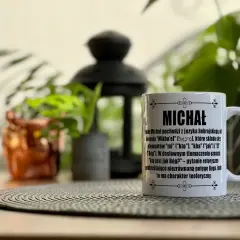 Biały kubek z napisem "MICHAŁ" i opisem znaczenia imienia, idealny na imieniny Michała. W tle rośliny i ozdobna latarenka.