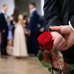 Elegancki mężczyzna w garniturze trzyma za plecami czerwoną różę, symboliczny gest na dzień mężczyzny w marcu. W tle widać bawiących się ludzi.