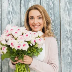 Uśmiechnięta kobieta z bukietem jasnoróżowych róż na tle drewnianej ściany.