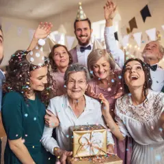 Radosna rodzina świętuje 50 rocznicę ślubu. Wszyscy w czapkach, sypią się konfetti. Babcia trzyma złoty prezent.