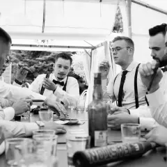 Panowie w eleganckich koszulach i muszkach świętują kawalerski przy stole z cygarami i butelką whisky.
