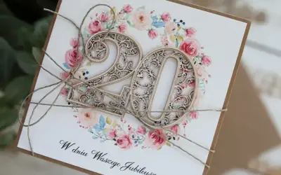 Elegancka kartka z napisem "W dniu Waszego Jubileuszu" i ażurowymi cyframi "20" na tle kwiatowej girlandy. Idealna na 20 rocznicę ślubu życzenia.