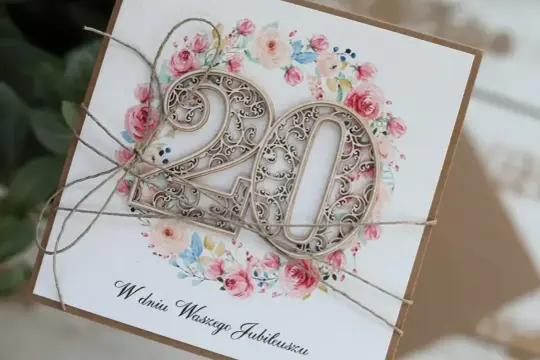 Elegancka kartka z napisem "W dniu Waszego Jubileuszu" i ażurowymi cyframi "20" na tle kwiatowej girlandy. Idealna na 20 rocznicę ślubu życzenia.