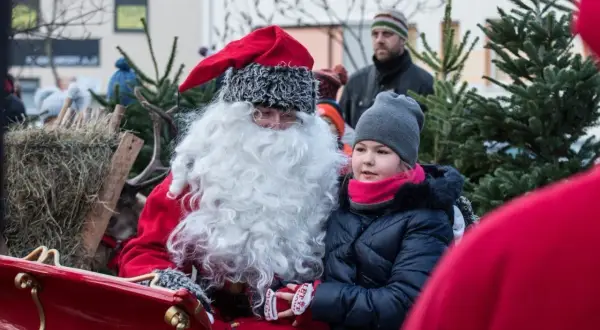 Święty Mikołaj w saniach z dziewczynką, która z zaciekawieniem patrzy przed siebie. W tle widać renifera i choinki.