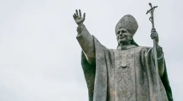 Pomnik Jana Pawła II z podniesioną ręką i krzyżem. Wierni odmawiają modlitwę do Jana Pawła II o uzdrowienie.