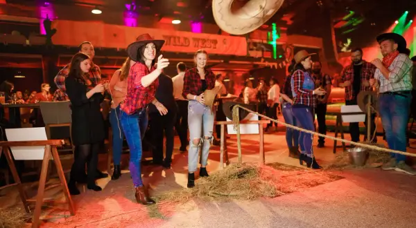 Dziewczyny w kapeluszach i kratkach bawią się na imprezie w stylu country. Rzucają kapeluszem, a w tle widać napis "Wild West".