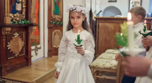 Dziewczynka w białej sukni i wianku trzyma zapaloną świecę. To ważny dzień, w którym klasa przystępuje do komunii.