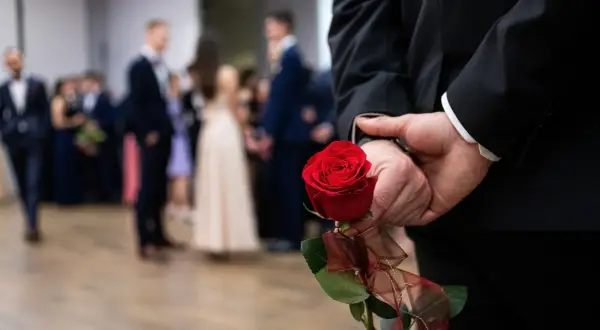 Elegancki mężczyzna w garniturze trzyma za plecami czerwoną różę, symboliczny gest na dzień mężczyzny w marcu. W tle widać bawiących się ludzi.