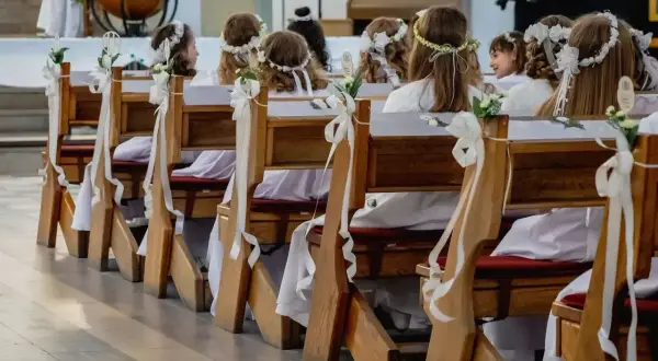 Dziewczynki w białych sukienkach i wiankach czekają na swoją pierwszą komunię świętą, siedząc w ławkach ozdobionych białymi wstążkami.