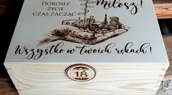 Drewniana skrzynka z grawerem na 18 urodziny od chrzestnej. Na wieczku napis "Dorosłe życie czas zacząć, Miłosz! Wszystko w Twoich rękach!" i grafika dłoni z symbolami podróży.