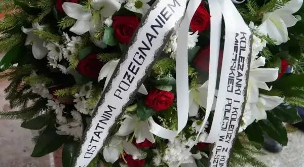 Wieniec z białych lilii, czerwonych róż i zieleni. Na białej szarfie widnieje oryginalny napis na szarfie pogrzebowej: "Z ostatnim pożegnaniem Koleżanki i Koledzy z Komendy Policji w Olsztynie".