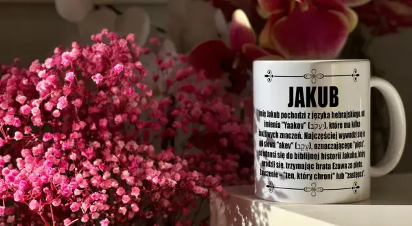 Kubek z napisem "JAKUB" i opisem imienia, otoczony delikatnymi, różowymi kwiatami. Idealny prezent na imieniny Jakuba.