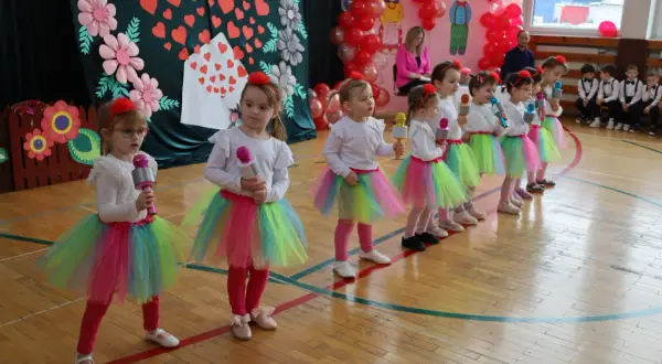 Dzieci w kolorowych spódniczkach śpiewają piosenkę na Dzień Dziecka, trzymając w rękach mikrofony.