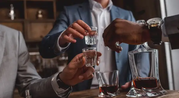 Mężczyźni w garniturach wznoszą kieliszki z alkoholem. W tle widać karafkę z whisky. To idealny moment na pępkowe!