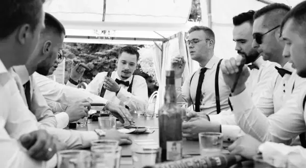 Panowie w eleganckich koszulach i muszkach świętują kawalerski przy stole z cygarami i butelką whisky.