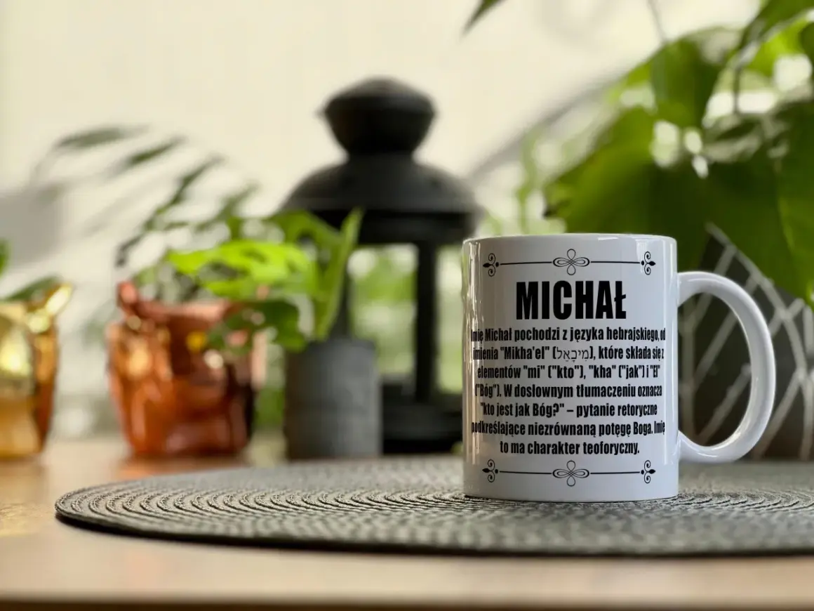 Biały kubek z napisem "MICHAŁ" i opisem znaczenia imienia, idealny na imieniny Michała. W tle rośliny i ozdobna latarenka.