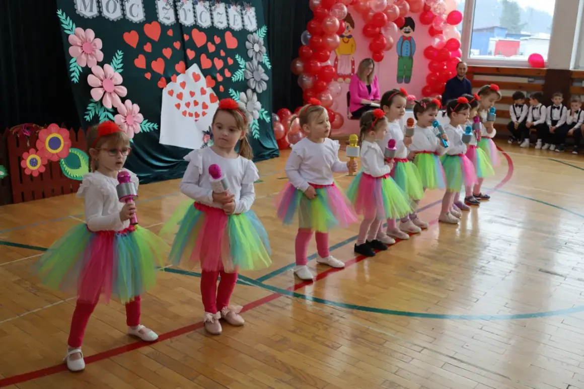 Dzieci w kolorowych spódniczkach śpiewają piosenkę na Dzień Dziecka, trzymając w rękach mikrofony.