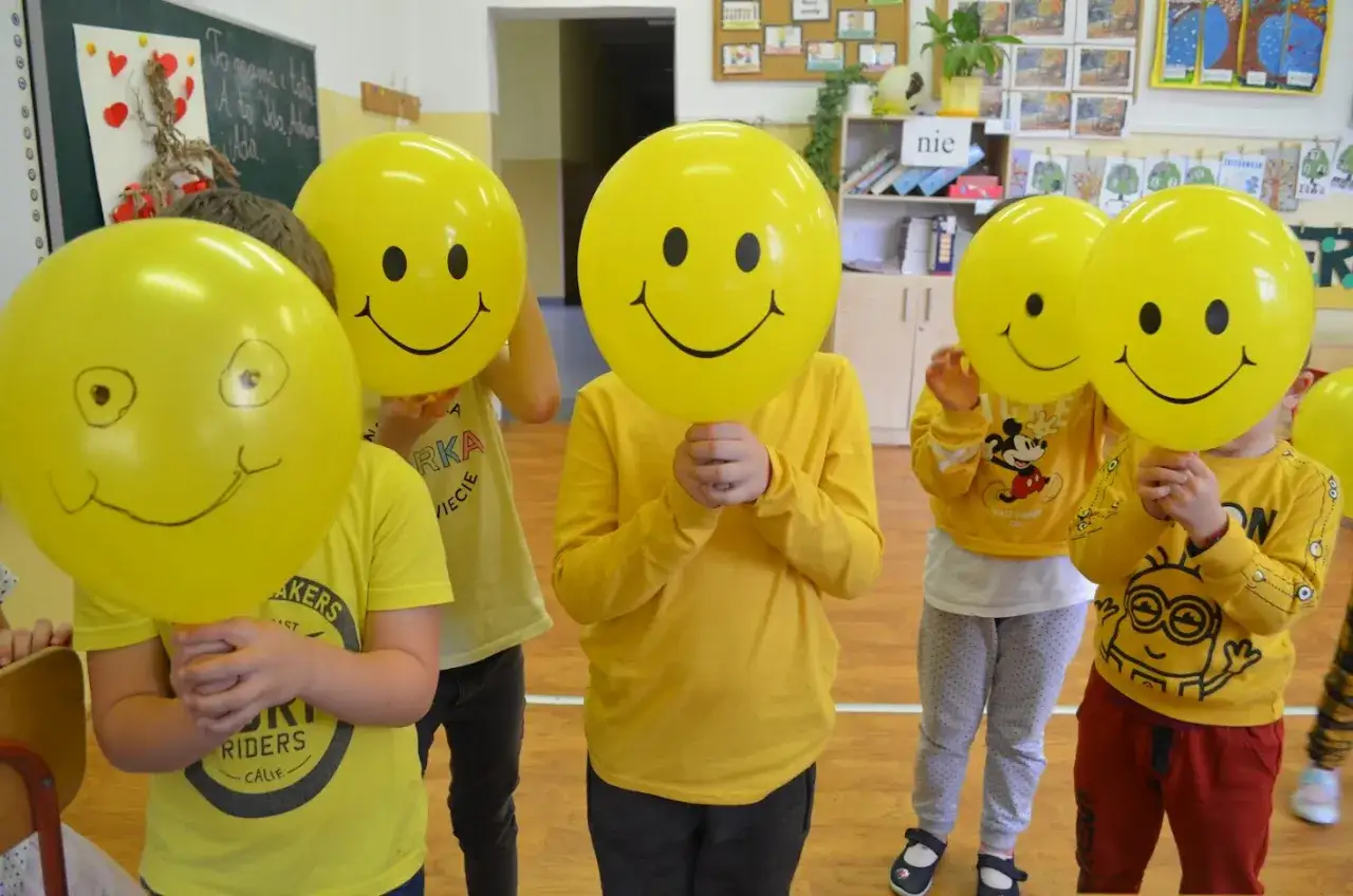 Dzieci w żółtych ubraniach trzymają balony ze śmiesznymi buźkami. To piękny dzień życzliwości w przedszkolu.