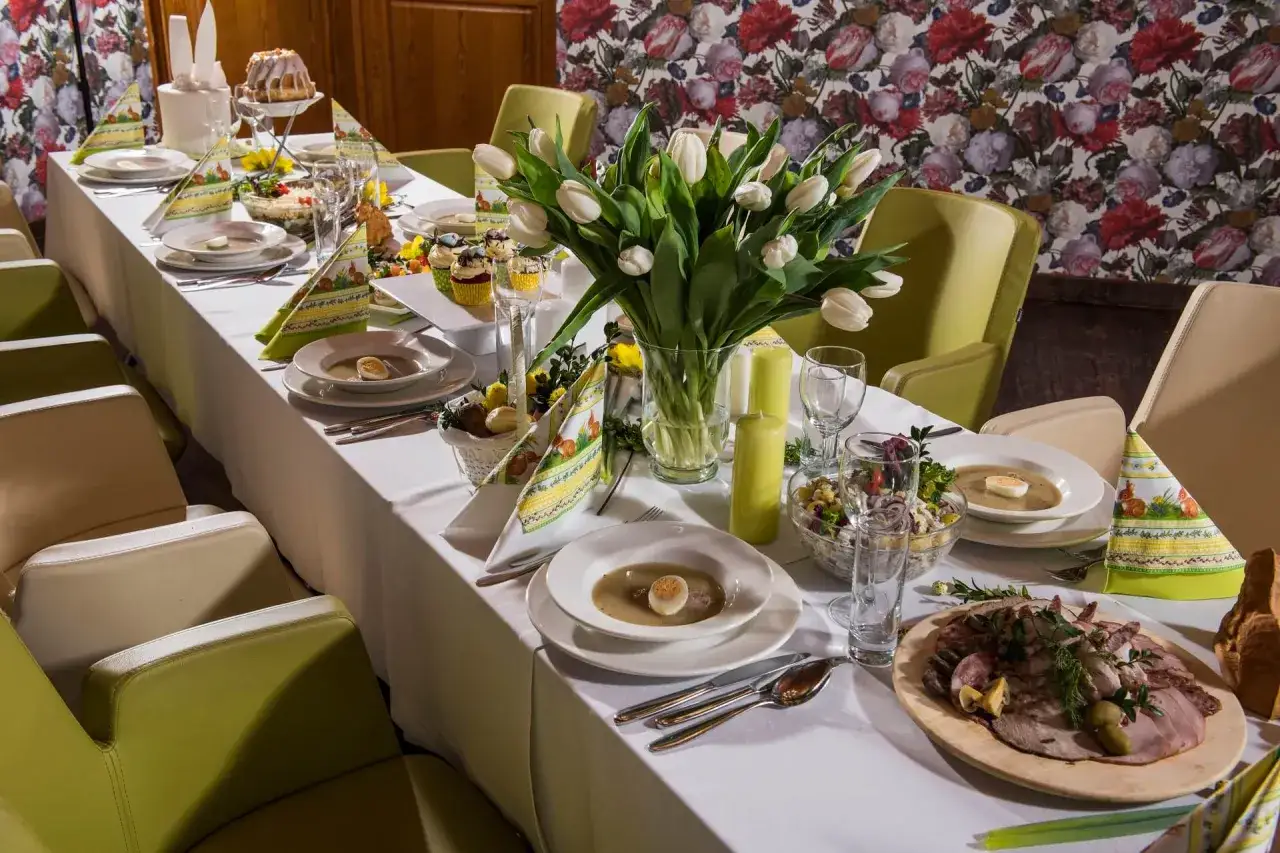 Uroczysty stół z potrawy wielkanocne lista: żurek, babka, babeczki, wędliny, sałatki. Białe tulipany i świece dodają elegancji.