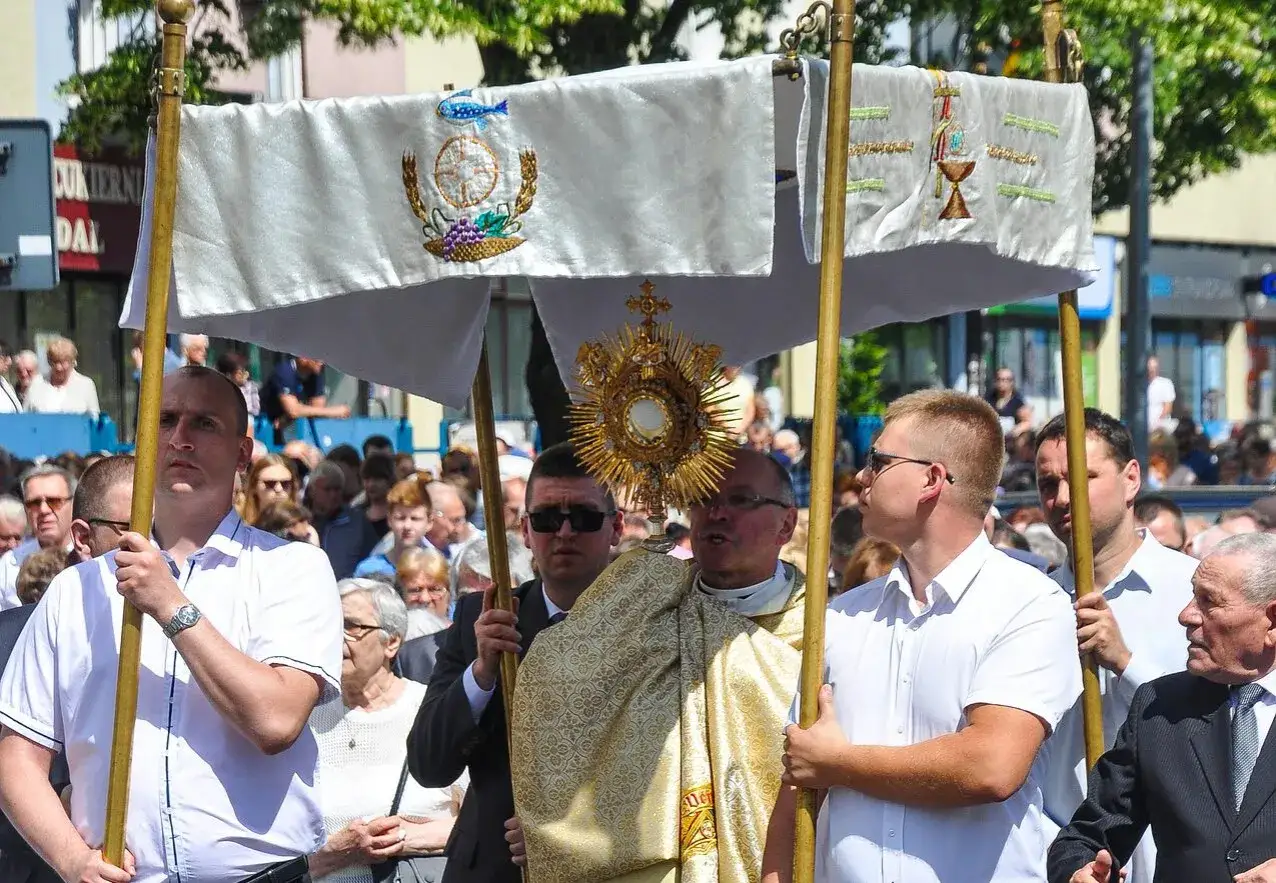 Procesja Bożego Ciała z monstrancją niesioną pod baldachimem. Ludzie idą ulicą, uczestnicząc w uroczystości.